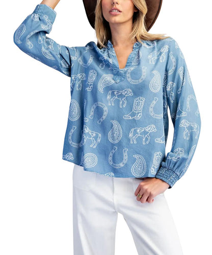 A woman wearing a light blue long-sleeve blouse with a white paisley and western-themed animal print, paired with white pants and a brown hat, against a plain white background.