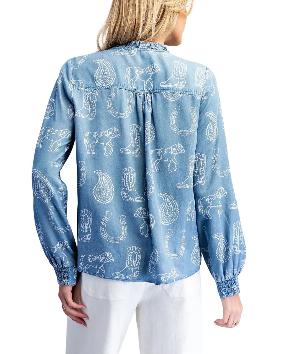 A woman shown from the back wearing a light blue long-sleeve button-up shirt with a white western-themed print of boots, horseshoes, and poodles, paired with white pants.