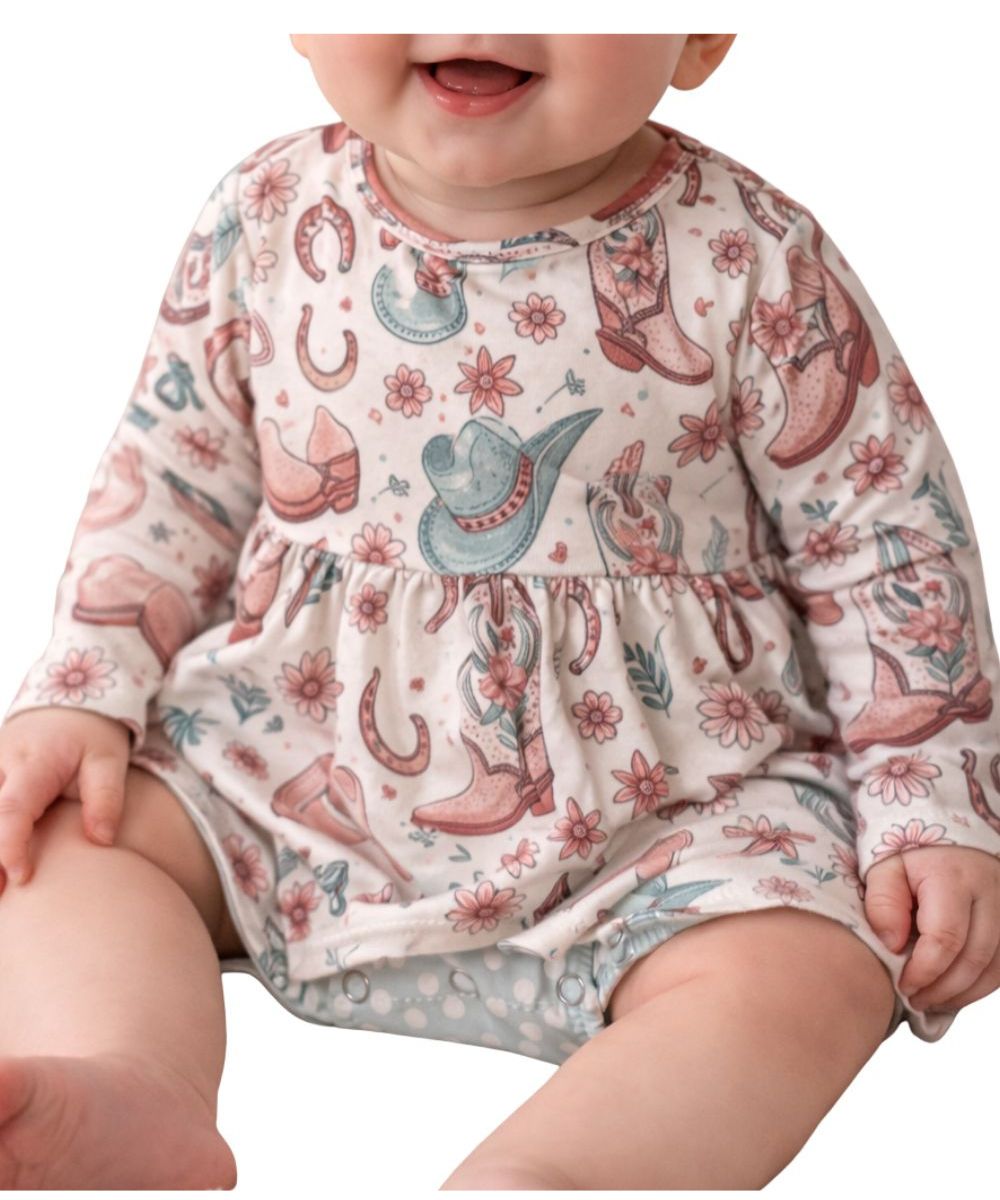 Baby girl's onesie with a soft cream base featuring a western-inspired print of cowboy boots, hats, horseshoes, and floral accents in muted pink and teal tones, designed with long sleeves and a gathered waist detail.